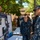 NMCSD NEC Fair U.S. Navy personnel are gathered at an outdoor career or rating fair, examining informational display boards. A female sailor wearing a name tag reading 'SUNK' stands behind a poster board titled 'Mass Communication Specialist (MC)', presenting information to fellow sailors. The event appears to take place in a courtyard of a military installation.