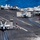 170612-N-BL637-125 The image shows a busy U.S. Navy aircraft carrier flight deck with multiple F/A-18 Hornet/Super Hornet jet aircraft positioned across the deck. Deck crew members in color-coded jerseys are performing various flight operations tasks. The carrier appears to be underway at sea, with deep blue ocean visible in the background.