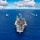 170612-N-BL637-012 An aerial photograph shows a naval carrier strike group underway in open blue ocean waters. A large aircraft carrier, bearing the hull number '70' on its bow, leads the formation with its flight deck loaded with multiple aircraft. Four escort vessels — appearing to be destroyers or cruisers — are positioned in a spread formation around the carrier.
