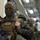 170614-N-GG458-071 A U.S. Marine in full combat gear, including MARPAT camouflage uniform, tactical vest, and face paint in a green/black camouflage pattern, stands in what appears to be an indoor staging area. He is holding an M4-style rifle and wearing name tape and unit insignia patches on his chest rig. Several other Marines in similar gear are visible in the background.
