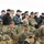 170614-N-PF515-006 The image shows a large group of multinational military personnel gathered together, likely observing a military exercise or demonstration. Multiple uniform types are visible, including US Marine Corps MARPAT camouflage, US Navy digital blue, and what appear to be Polish and other NATO allied uniforms. Personnel are positioned both behind a camouflage netting barrier in the foreground and standing in a crowd in the background.