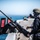 170613-N-TV230-037 A U.S. Navy sailor in full combat gear stands on the deck of a naval vessel operating on open water. The sailor is positioned at a mounted crew-served weapon, appearing to conduct a readiness check or security watch. The image is taken in bright daylight with a clear blue sky and sea visible in the background.