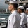 170614-N-LK571-135 U.S. Navy sailors in dress white uniforms stand at attention on the deck of a naval vessel, appearing to render honors as they pass the USS Arizona Memorial at Pearl Harbor, Hawaii. The white memorial structure is clearly visible in the harbor background, with the city of Honolulu and the Ko'olau mountain range visible in the distance. At least one officer in a khaki uniform with gold insignia is also present among the enlisted sailors.