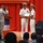 Commander, Amphibious Force 7th Fleet awards Capt. Paradise at Blue Ridge's change of command ceremony. A formal military ceremony is taking place on a stage with a red curtain backdrop. Two naval officers in white dress uniforms are shaking hands while jointly holding what appears to be a certificate or award. A third uniformed individual is photographing the moment while an audience watches from the foreground.