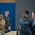 170615-N-SD711-0154 The image shows a group of people in an indoor facility, likely a gymnasium or community center, engaged in conversation. Two U.S. Navy personnel in digital camouflage uniforms (NWU Type I) are interacting with civilians, including a woman in a green cardigan and a man in a dark business suit. The setting appears to be an official or community engagement event.