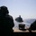 170609-N-FM530-005 Two silhouetted naval personnel are observing a military vessel from the deck of another ship, likely during an underway replenishment or formation sailing exercise. The observer on the left is wearing communication headgear with visible text 'LPD 19' on the helmet. The scene is set on open ocean under bright, hazy sunlight.