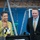 170615-N-SD711-0157 Two individuals stand together in what appears to be a gymnasium or sports facility. A woman in a yellow-green cardigan and patterned blouse is smiling and gesturing, while a man in a dark suit with a gold striped tie stands beside her. Camera equipment is visible in the foreground, suggesting a media or broadcast setting.