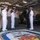 170613-N-FM530-028 A group of military personnel and one civilian are gathered in what appears to be the vehicle/hangar deck of the USS Mesa Verde, a U.S. Navy amphibious transport dock ship. The group includes U.S. Navy sailors in white dress uniforms and U.S. Marines in olive drab service uniforms, along with a civilian in a dark suit. They appear to be engaged in a formal briefing or official visit, standing near military vehicles and equipment.
