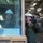 170614-N-LC424-0745 Two military personnel wearing white hard hats and olive drab uniforms are visible in what appears to be the interior deck of a naval vessel or large military facility. One individual is seated inside a vehicle cab visible through a glass window, while the other stands outside pointing or gesturing with a gloved hand. The setting suggests a vehicle staging or maintenance area aboard a ship.
