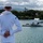 170614-N-BL637-068 A U.S. Navy sailor in dress whites stands with hands clasped behind their back, facing the USS Arizona Memorial at Pearl Harbor, Hawaii. The white memorial structure is visible over the sunken battleship, with 'USS ARIZONA BB 39' marked on a buoy nearby. An American flag flies above the memorial against a partly cloudy sky with green tropical vegetation and mountains in the background.