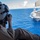 170612-N-YA681-008 A military aircrew member in a flight suit and helmet is photographing a large aircraft carrier from the open door of a helicopter over open ocean. The photographer is seated at the edge of the helicopter bay with legs dangling, capturing the carrier from an elevated aerial perspective. The scene depicts a routine aerial reconnaissance or documentation mission over blue open water.