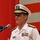 Commander, Amphibious Force 7th Fleet gives remarks at Blue Ridge's change of command ceremony. A U.S. Navy officer in white dress uniform is speaking at a podium microphone during what appears to be an official military ceremony or event. The officer wears a combination cover with gold scrambled eggs indicating flag or senior officer rank, along with multiple service ribbons and a name tag. A red and white background, possibly a flag or banner, is visible behind him.