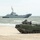 170614-N-PF515-011 A military amphibious assault vehicle (AAV) is positioned on a sandy beach with a naval warship visible offshore in the background. The scene appears to depict an amphibious landing exercise or military operation, with at least one soldier visible in the open hatch of the tracked vehicle. The overcast sky and choppy sea conditions suggest a northern or temperate coastal environment.
