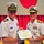 Commander, Amphibious Force 7th Fleet awards Capt. Paradise at Blue Ridge's change of command ceremony. Two U.S. Navy officers in white dress uniforms are jointly holding a Legion of Merit certificate issued by The United States of America. The ceremony appears to be taking place in a formal indoor venue decorated with red curtains and what appears to be a Japanese flag in the background. A third uniformed individual is partially visible in the background.