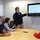 170607-N-HN707-363 A presenter is standing and pointing to a wall-mounted Toshiba display screen showing a slide titled 'Background: EpiData Center Department' in a conference room setting. Three seated individuals around a round table are listening and engaging with the presentation. The content appears to be related to Navy and Marine Corps Public Health Center suicide prevention epidemiological research.