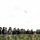 170614-N-PF515-005 A group of military personnel and civilians in various uniforms are observing a large military aircraft flying overhead. The observers are standing behind a camouflage-netted barrier, suggesting this is a formal military demonstration or exercise review. Multiple national uniforms are visible, indicating a multinational or NATO-affiliated event.