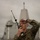170614-N-GG458-144 A U.S. Marine in MARPAT digital camouflage uniform is using a handheld military radio (likely a PRC-152 or similar tactical radio) while shielding his eyes to observe something in the distance. He is positioned on what appears to be the deck of a U.S. Navy vessel, with a large warship — identifiable by its angular, stealth-design superstructure consistent with a Zumwalt-class destroyer or Littoral Combat Ship — visible in the background. The overcast sky and maritime setting suggest an active naval operation or exercise.