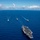 170612-N-YA681-006 An aerial photograph shows a U.S. Navy carrier strike group underway on open ocean. A large aircraft carrier dominates the foreground with its flight deck loaded with aircraft, while three escort vessels — likely guided-missile cruisers or destroyers — are positioned ahead in a triangular formation. All vessels are leaving visible wakes as they transit through calm, deep-blue waters under a partly cloudy sky.