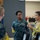 170615-N-SD711-0141 The image shows a group of individuals in what appears to be a military facility or training room. A U.S. Navy sailor in digital camouflage uniform (NWU) with the name tape 'MOORE' is engaged in conversation with a civilian woman wearing a yellow cardigan. Two other uniformed personnel are visible in the foreground with their backs to the camera.