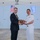 170613-N-FM530-122 Two individuals are posing together while jointly holding a wooden commemorative plaque during what appears to be an official ceremony. One person is wearing a dark business suit with a yellow tie, while the other is dressed in a white naval officer's uniform with rank insignia. The setting appears to be aboard a naval vessel, with both an American flag and a Greek flag visible in the background.