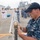 Sterett-Dewey Surface Action Group Deployment A U.S. Navy sailor in digital camouflage uniform is working with rope/line on a metal pole or stanchion at a naval pier. In the background, groups of military personnel in different uniforms — including white dress uniforms and blue camouflage — are gathered, suggesting a multinational or joint naval event. Several warships, including one marked '885', are visible docked at the pier.