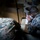 Two military personnel in MARPAT (Marine pattern) camouflage uniforms are crouched on the floor examining a document or map. A third individual in the same uniform is visible in the upper right corner, standing. The scene appears to be inside a building, with the two primary subjects focused on reviewing written materials under limited lighting.