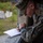 A male service member in US Marine Corps MARPAT digital camouflage uniform is leaning over a concrete ledge or wall, writing on a white sheet of paper with a pen. He is wearing a camouflage cover (cap) and a tactical wristwatch. The background shows an outdoor environment with greenery and what appears to be a training or base facility.