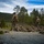 A group of military personnel in camouflage uniforms are conducting what appears to be a live-fire or tactical training exercise in an outdoor range setting. Several soldiers are in the prone position along a gravel firing line, while two standing individuals appear to be instructors or range safety officers overseeing the exercise. The setting is a forested, hilly landscape consistent with Scandinavian or Northern European terrain.