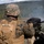 A U.S. Marine identified by the name tape 'ANDREWS' is operating a crew-served automatic grenade launcher, likely an Mk 19, during what appears to be a live-fire training exercise. The Marine is wearing MARPAT digital camouflage utilities, a Lightweight Helmet (LWH), and a tan tactical vest with MOLLE webbing. Smoke is visible downrange, consistent with recent weapons discharge, set against a forested valley landscape.