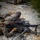 U.S. Marines in MARPAT camouflage uniforms are conducting a live-fire training exercise in a prone position on a gravel surface. The foreground shows two Marines operating what appears to be an M240 machine gun mounted on a bipod, with numerous spent brass casings scattered around the weapon. Additional personnel and weapons are visible in the background, indicating a squad-level firing line exercise.
