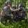 Three military personnel in MARPAT (Marine pattern) camouflage uniforms are crouched in a forested area, with one soldier pointing in a direction while holding a radio handset. The scene appears to be a field training exercise or tactical operation in a woodland environment. Additional personnel are visible in the background moving through the vegetation.