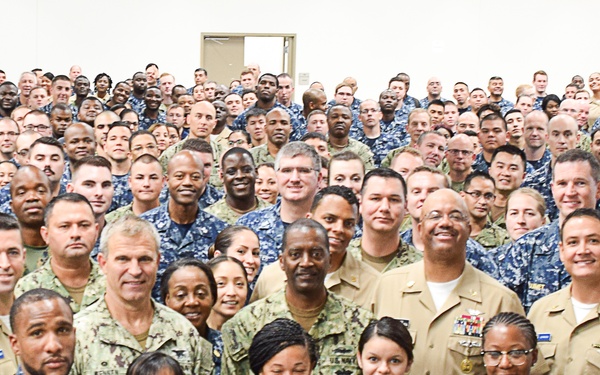 A large group photograph of U.S. Navy military personnel posing together in what appears to be an indoor facility or auditorium. The group consists of hundreds of service members wearing various Navy uniforms including the Navy Working Uniform (NWU) digital camouflage pattern and khaki/tan uniforms. The diverse group includes both male and female sailors of various ranks.
