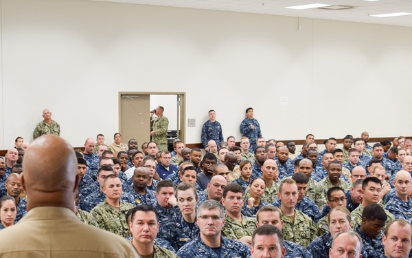 A senior military officer in tan/khaki service uniform addresses a large assembly of U.S. Navy personnel seated in rows of chairs in a large meeting hall. The audience consists of hundreds of sailors wearing Navy Working Uniform (NWU) blue digital camouflage, along with some personnel in tan/desert camouflage. The setting appears to be an all-hands call or command briefing at a naval installation.