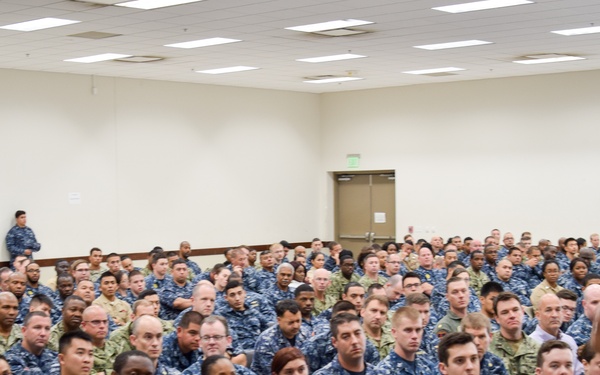 A senior U.S. Navy officer in khaki service uniform addresses a large assembly of Navy personnel seated in a conference or meeting hall. The audience consists of hundreds of sailors wearing Navy Working Uniform (NWU) blue digital camouflage pattern, along with some personnel in tan/green camouflage uniforms. The setting appears to be an all-hands call or command briefing at a military installation.