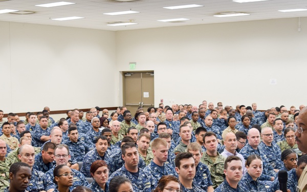 A senior military officer in tan/khaki service uniform is addressing a large assembly of Navy personnel seated in rows in a conference or auditorium-style room. The majority of attendees are wearing Navy Working Uniform (NWU) digital blue camouflage, with some individuals in olive/green military uniforms. The speaker is holding a microphone and appears to be conducting an all-hands briefing or command presentation.