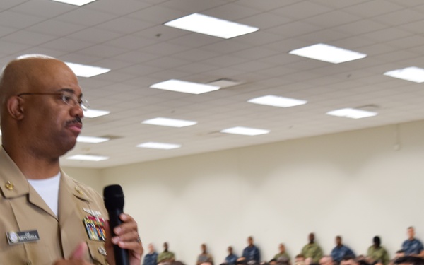 A senior U.S. Navy officer in khaki service uniform is addressing a large audience of military personnel in a briefing room or auditorium. The speaker is holding a microphone and appears to be delivering a speech or all-hands briefing to dozens of sailors seated in rows. The audience is wearing a mix of Navy Working Uniform (NWU) blue digital camouflage and tan/green camouflage uniforms.
