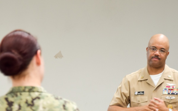 Two U.S. Navy personnel are engaged in what appears to be a briefing or instructional session indoors. A senior officer in Service Khaki uniform with multiple ribbons and insignia is speaking or presenting, while a junior sailor in Navy Working Uniform (NWU) camouflage pattern faces them with their back to the camera. The setting appears to be a conference room or classroom environment.