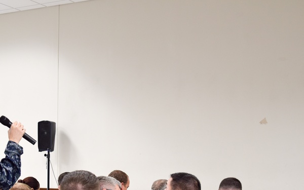A military briefing or town hall meeting is taking place in an indoor facility. A female sailor in Navy Working Uniform (NWU) is standing and speaking into a microphone, while a senior officer in khaki service uniform listens from the right side. Multiple sailors seated in folding chairs are turned to observe the exchange.