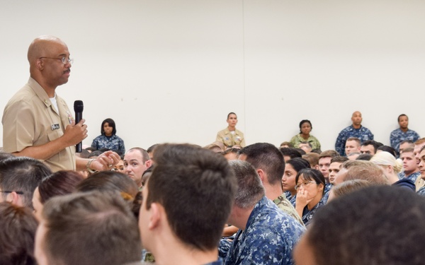 A senior military officer in tan/khaki uniform is addressing a large group of Navy personnel seated on the floor in what appears to be a briefing or all-hands meeting. The speaker is holding a microphone and gesturing while speaking to an audience of dozens of sailors wearing Navy Working Uniform (NWU) blue digital camouflage. The setting appears to be a large indoor facility such as a gymnasium or hangar bay.