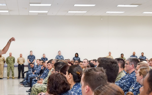 A senior U.S. Navy officer in tan service uniform is addressing a large group of sailors wearing Navy Working Uniform (NWU) blue digital camouflage. The speaker is holding a microphone and gesturing expressively to an audience seated on the floor and standing along the back wall. The setting appears to be a large indoor military facility, likely a briefing hall or gymnasium.
