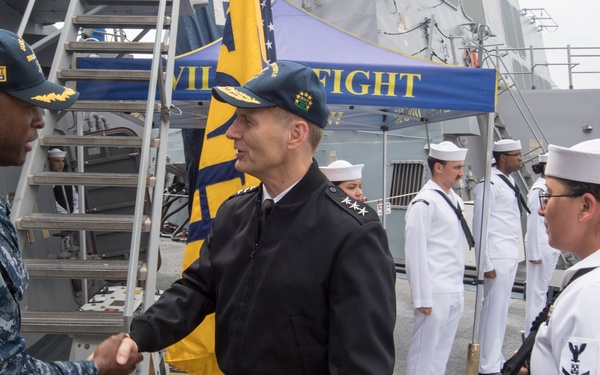 A formal military ceremony is taking place on the deck of a naval vessel, likely a U.S. Navy ship. A senior officer in a dark black uniform with gold insignia is shaking hands with a U.S. Navy sailor in digital camouflage (NWU) while a line of sailors in dress white uniforms stands at attention in the background. A banner reading 'FIGHT' in blue and gold is visible, suggesting a unit or command motto display.