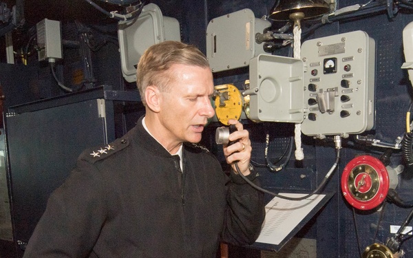 A senior naval officer in a dark dress uniform is standing in what appears to be a ship's bridge or control room, speaking into a handheld communication device. He wears shoulder insignia consistent with a high-ranking officer (three stars visible). The setting contains various nautical equipment, fire safety apparatus, and control panels mounted on dark blue bulkheads.