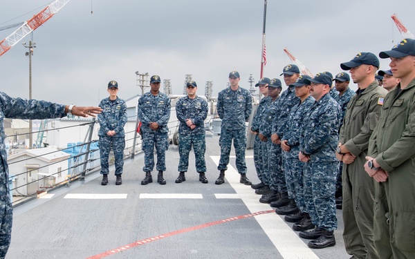A senior naval officer in black service dress uniform addresses a formation of sailors on what appears to be a ship's flight deck or pier. The personnel are divided into two visible uniform groups: those in Navy Working Uniform (NWU) digital camouflage and those in olive drab flight suits. The setting appears to be a naval installation, likely in Asia based on the visible cranes and infrastructure in the background.