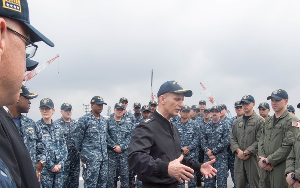 A senior naval officer in a black dress uniform is addressing a large group of U.S. Navy personnel on what appears to be the deck of a naval vessel. The officer is gesturing with both hands while speaking to sailors dressed in Navy Working Uniform (NWU) digital camouflage and some in olive green flight suits. The setting appears to be an outdoor deck environment with an overcast sky in the background.