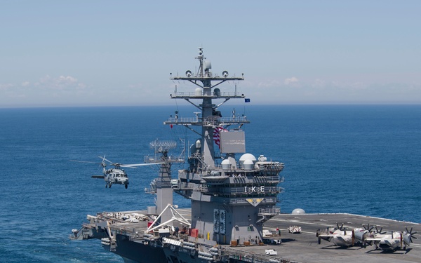 An aerial photograph of a U.S. Navy aircraft carrier underway at sea, identifiable by the letters 'IKE' on the island superstructure, indicating USS Dwight D. Eisenhower (CVN-69). The flight deck is populated with fixed-wing aircraft and a helicopter is visible in flight alongside the island. The vessel is operating in open blue ocean under clear skies.