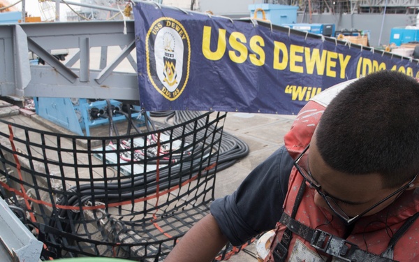 A Navy sailor wearing a red life vest and glasses is performing maintenance or mechanical work on deck equipment aboard a naval vessel. A banner in the background identifies the ship as the USS Dewey (DDG-105). The sailor is holding a cup containing what appears to be paint or a liquid substance while working with a tool on a fitting or cleat.