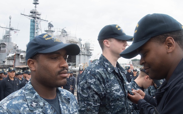 A military ceremony is taking place on what appears to be a naval vessel or pier, where a service member in a dark navy uniform is pinning a medal or insignia onto the uniform of another sailor wearing Navy Working Uniform (NWU) digital camouflage. The recipient, whose name tag reads 'RAY,' stands at attention while the presentation is made. A large formation of Navy personnel in dark uniforms stands in the background, with naval ships visible in the distance.