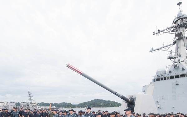 A U.S. Navy officer addresses a large formation of sailors on the deck of a warship, likely an Arleigh Burke-class guided-missile destroyer, moored at a naval base. The ceremony appears to be a formal all-hands or awards event, with sailors standing in organized ranks facing the speaker at a podium. The setting appears to be a forward-deployed naval installation, possibly Yokosuka, Japan, based on the surrounding harbor and terrain.