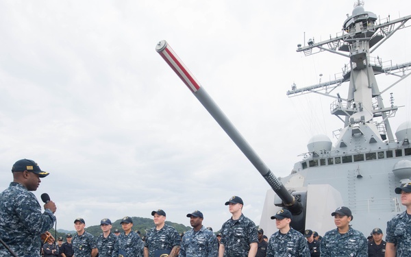 A U.S. Navy ceremony is taking place on the deck of a naval warship, likely a destroyer or cruiser, with a large gun barrel prominently visible in the background. A senior officer is addressing a group of sailors standing in formation, several of whom are holding blue folders or certificates, suggesting an awards or recognition ceremony. A large crowd of additional naval personnel observes from the sides.