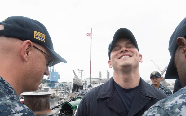 Three U.S. Navy personnel are gathered on what appears to be a naval pier or dock, with one sailor in the center smiling broadly while two others in digital camouflage uniforms (NWU Type I) appear to be pinning something on him, suggesting a promotion or award ceremony. The background shows naval vessels, industrial dock equipment, and an American flag on a flagpole. The scene conveys a positive, celebratory military moment.